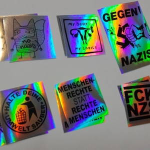 Könnte beinhalten: Eine Sammlung holografischer Aufkleber mit verschiedenen Designs und Texten. Ein Aufkleber zeigt eine Fuchsgrafik mit dem Text "NO WARS". Weitere Aufkleber enthalten Sätze wie "My Body My Choice", "GEGEN NAZIS" und "HALTE DEINE UMWELT SAUBER".