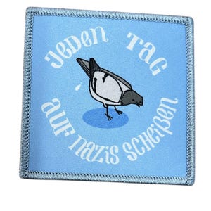 Könnte beinhalten: Ein quadratischer Aufnäher mit hellblauem Hintergrund und gesticktem Rand. Der Aufnäher zeigt eine Cartoon-Vogel-Illustration und deutschen Text, der lautet: "Jeden Tag auf Nazis scheißen."