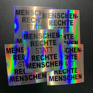 Könnte beinhalten: Ein Satz von fünf schillernden holografischen Aufklebern mit dem Text "MENSCHEN RECHTE STATT RECHTE MENSCHEN" in schwarzer Schrift.