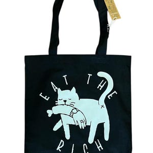 Könnte beinhalten: Schwarze Tragetasche mit weißer Grafik einer Katze, die einen Fisch hält, mit den Worten "EAT THE RICH" in Weiß. Die Tasche hat schwarze Griffe und ein angebrachtes Etikett.