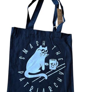 Könnte beinhalten: Marineblaue Tragetasche mit einer weißen Grafik einer Katze mit Sonnenbrille und einer Tasse mit dem Männersymbol. Der Text "SMASH THE PATRIARCHY" ist in einem Kreis um die Katze angeordnet.