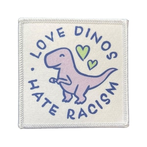 Könnte beinhalten: Quadratischer Aufnäher mit weißem Hintergrund und genähter Umrandung. Der Aufnäher zeigt eine lila Dinosaurier-Illustration mit dem Text "LOVE DINOS HATE RACISM" in Blau sowie grüne Herzen.