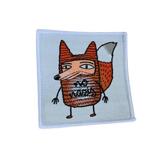 Könnte beinhalten: Ein weißer Stoff-Patch mit einem Cartoon-Fuchs, der eine rote und orangefarbene Strickmütze und einen Strickpullover trägt. Der Fuchs hat eine schwarze Nase und Augen und einen weißen Schwanz. Der Pullover hat die Worte "We are animals" darauf geschrieben.