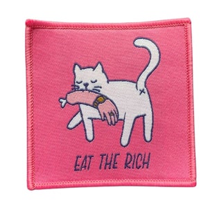 Könnte beinhalten: Rosa gestickter Patch mit einer weißen Katze, die einen rosafarbenen menschlichen Arm frisst. Der Text "EAT THE RICH" befindet sich unter der Katze.