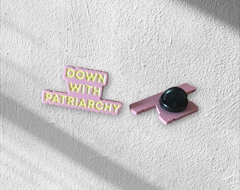 May include: A pink enamel pin with the text "DOWN WITH PATRIARCHY" in yellow lettering. The pin is a rectangular shape with a black pin back. The pin is on a white textured surface.