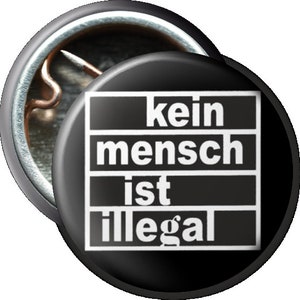 Könnte beinhalten: Schwarz-weißer Knopf mit dem Text "kein mensch ist illegal" in Weiß.