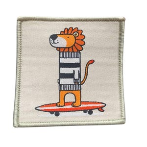 Könnte beinhalten: Ein beigefarbener Stoff-Patch mit einem Löwen, der einen gestreiften Pullover trägt und auf einem Surfbrett reitet. Der Löwe hat orangefarbenes Fell und eine Mähne, und das Surfbrett ist rot mit weißen Streifen.
