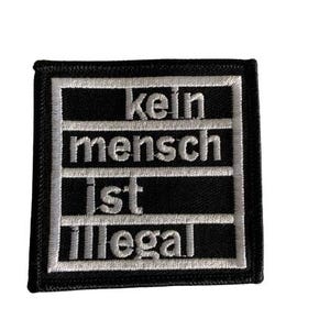 Könnte beinhalten: Schwarz-weißer, gestickter Aufnäher mit dem deutschen Text „kein mensch ist illegal“ (kein Mensch ist illegal). Der Aufnäher hat eine rechteckige Form mit schwarzem Rand und weißen Nähten.