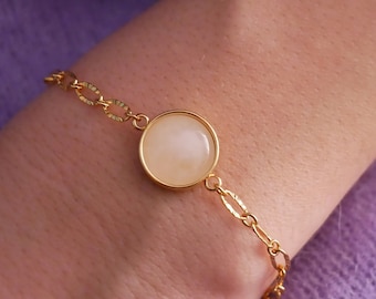 Puede incluir: Pulsera dorada con una piedra grande y redonda de color crema engastada en un bisel. La pulsera tiene una delicada cadena con eslabones ovalados. La joya se muestra en la muñeca de una persona sobre un fondo morado suave.