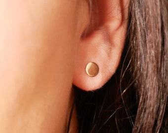 May include: A close-up of a person's ear with a small, round, gold earring. The earring is a simple, minimalist design.
