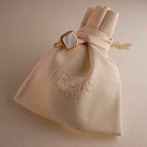 May include: A white and gold ring with a square-shaped mother-of-pearl stone. The ring is in a white velvet pouch with the word "IRBIS PARIS" printed on it.