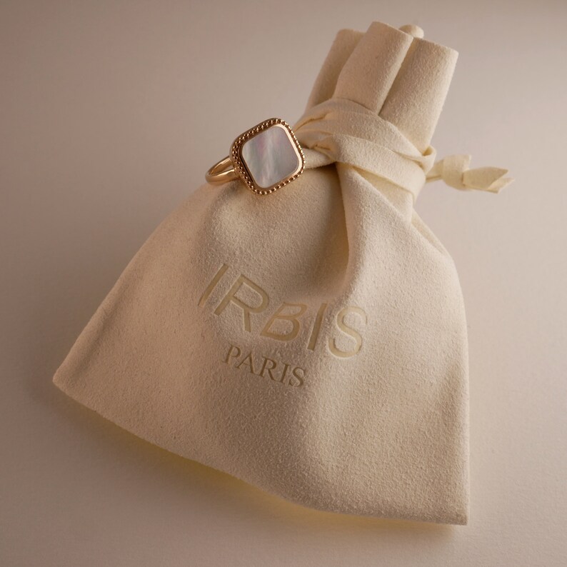 May include: A white and gold ring with a square-shaped mother-of-pearl stone. The ring is in a white velvet pouch with the word "IRBIS PARIS" printed on it.