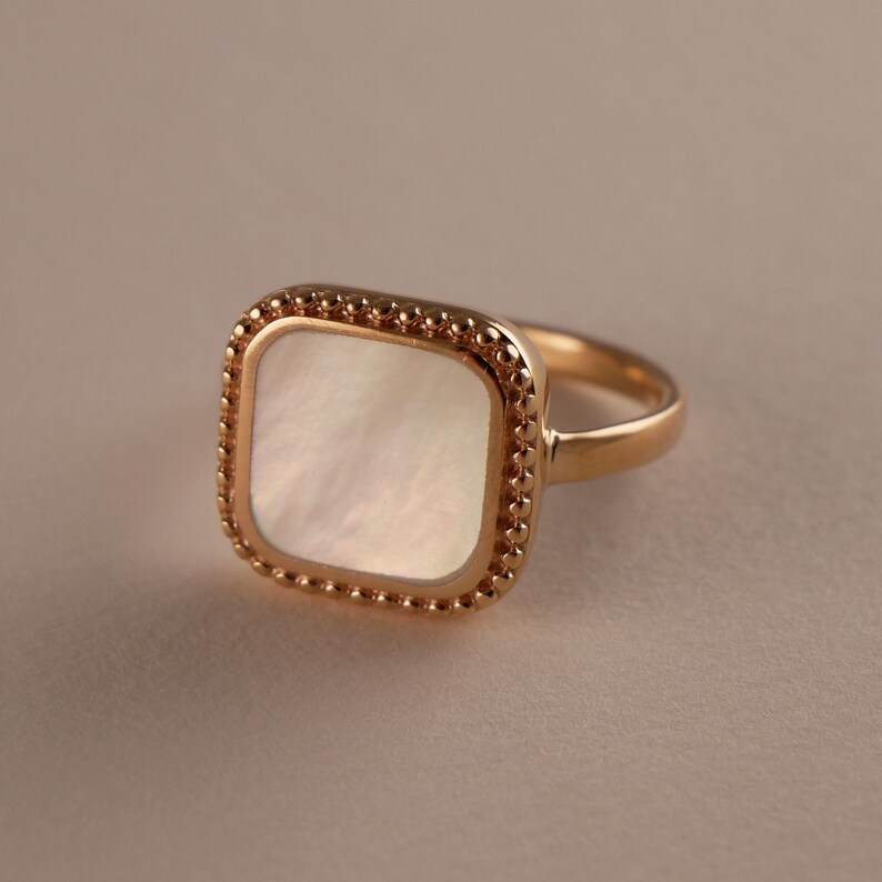 May include: A gold ring with a square-shaped mother-of-pearl stone set in a beaded bezel.