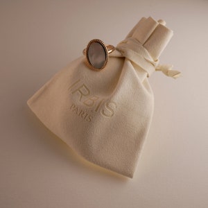 May include: A white velvet jewelry pouch with the word "IRBIS PARIS" embossed on it. A gold ring with a white oval stone is sitting on top of the pouch.