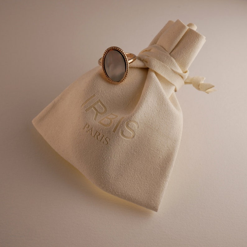 May include: A white velvet jewelry pouch with the word "IRBIS PARIS" embossed on it. A gold ring with a white oval stone is sitting on top of the pouch.