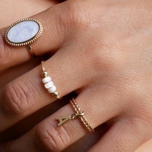May include: A hand wearing three gold rings. The first ring has a large oval white stone with a gold border. The second ring has a row of small white beads. The third ring has a gold chain with a letter A charm.