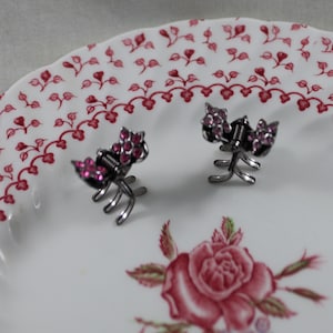 May include: Two silver hair clips with pink rhinestones on a white plate with a red floral pattern.