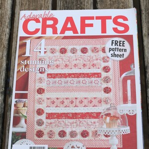 May include: A magazine cover featuring a red and white quilt with a floral pattern. The magazine is titled "Adorable Crafts" and has the number "14" on the cover. The text "FREE pattern sheet" is also on the cover. The magazine is on a wooden surface.