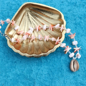 May include: A pink and white shell necklace with a cowrie shell charm, displayed on a gold seashell dish.