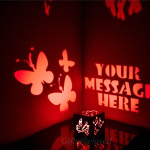 Könnte beinhalten: Eine schwarze Holzkiste mit einem Licht im Inneren wirft einen roten Schatten an die Wand. Der Schatten enthält Schmetterlinge und den Text "YOUR MESSAGE HERE".