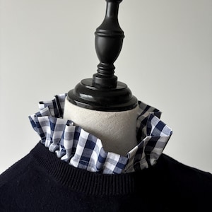 Op de afbeelding: Een marineblauwe trui met een afneembare blauw-witte geruite kraag. De kraag is gerimpeld en voegt een decoratief element toe aan de trui. De trui wordt getoond op een zwarte mannequin.