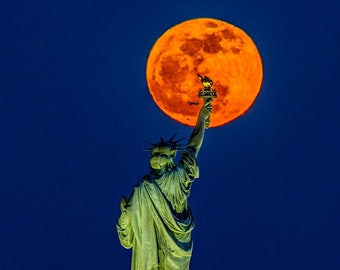 May include: The Statue of Liberty stands tall against a vibrant orange full moon in a dark blue sky. The statue is illuminated, highlighting its green copper color. The moon is a bright orange, almost red, and fills the upper portion of the image.