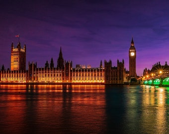 May include: A night view of the Houses of Parliament and Big Ben in London, England. The buildings are illuminated with warm yellow lights, and the river Thames is reflecting the lights. A bridge is in the foreground.