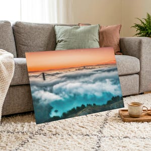 May include: A rectangular wall art print featuring a landscape photograph of the Golden Gate Bridge partially obscured by a thick layer of fog, with a vibrant orange sunset sky above and dark green hills visible in the foreground below the clouds.