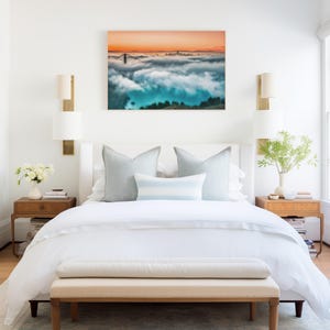 May include: A bedroom featuring a white bed with light blue pillows, two wooden nightstands with lamps, and a rectangular wall art print depicting a bridge and city skyline emerging from thick clouds under an orange sunset sky.