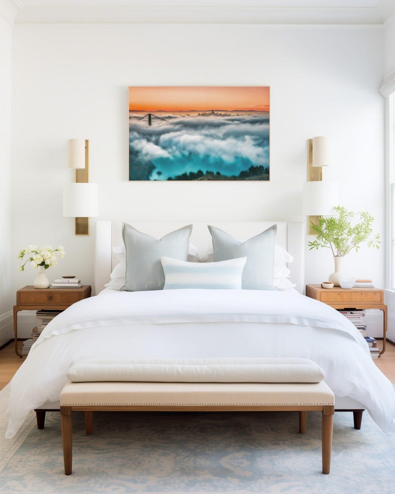 May include: A bedroom featuring a white bed with light blue pillows, two wooden nightstands with lamps, and a rectangular wall art print depicting a bridge and city skyline emerging from thick clouds under an orange sunset sky.
