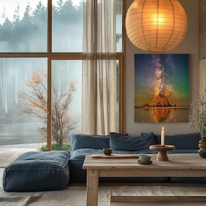 May include: A rectangular wall art print featuring a vibrant night sky with a colourful nebula and stars above a rocky coastal formation. The artwork is displayed on a wall above a blue sofa, next to a large window, a wooden table, and a paper lantern.