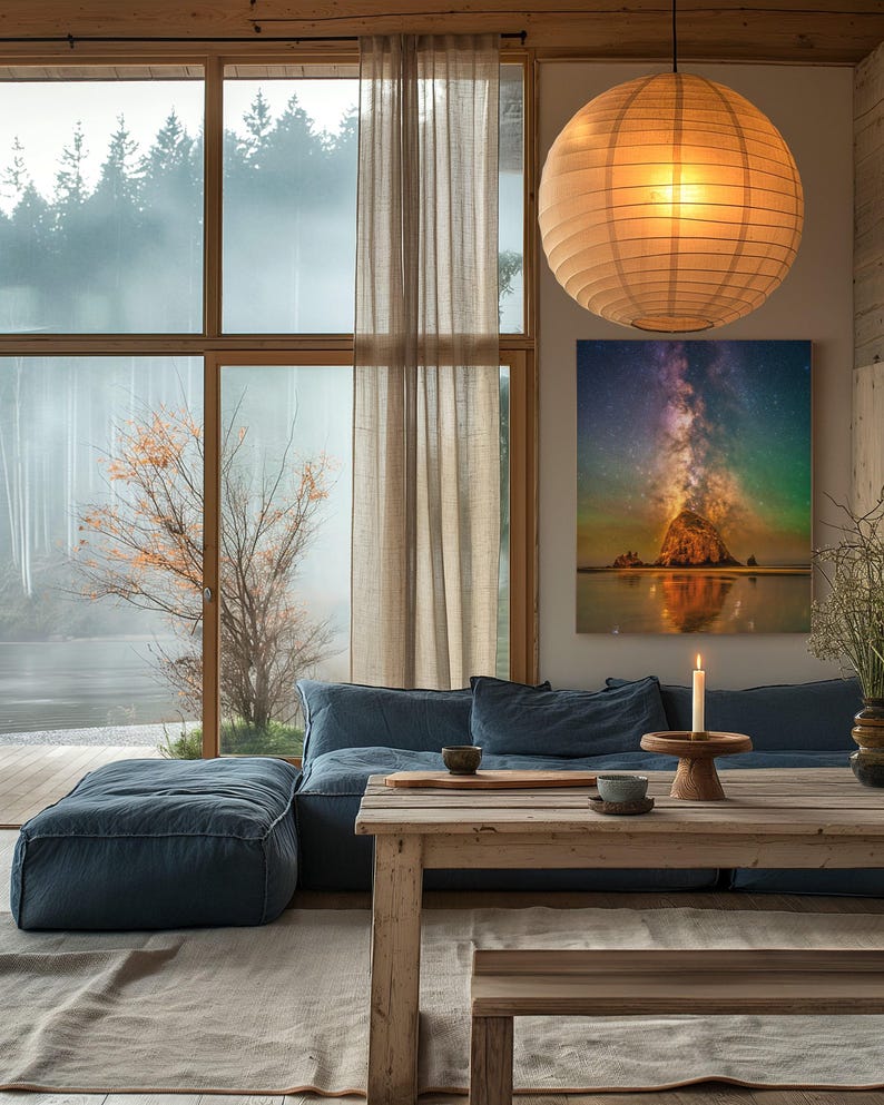 May include: A rectangular wall art print featuring a vibrant night sky with a colourful nebula and stars above a rocky coastal formation. The artwork is displayed on a wall above a blue sofa, next to a large window, a wooden table, and a paper lantern.