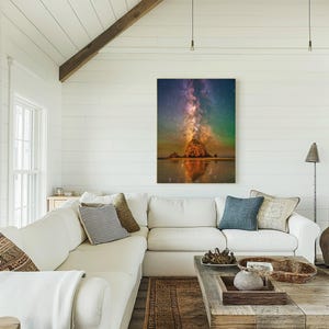 May include: A vertical wall art print featuring a night sky filled with a vibrant purple and orange Milky Way galaxy above a large, dark rock formation on a beach, with the scene reflected in the wet sand below the rock at the water's edge.