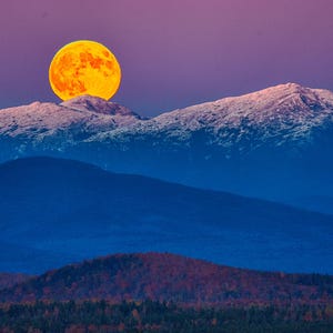 Op de afbeelding: Een grote, feloranje maan komt op boven een besneeuwde bergketen. De lucht is een levendig paars en roze.