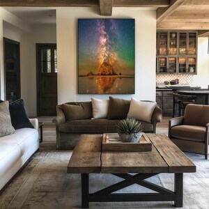 May include: A large rectangular wall art print featuring a vibrant night sky with a glowing Milky Way galaxy above a dark coastal rock formation, reflecting onto the wet sand below, displayed in a frame on a white wall above a brown sofa.