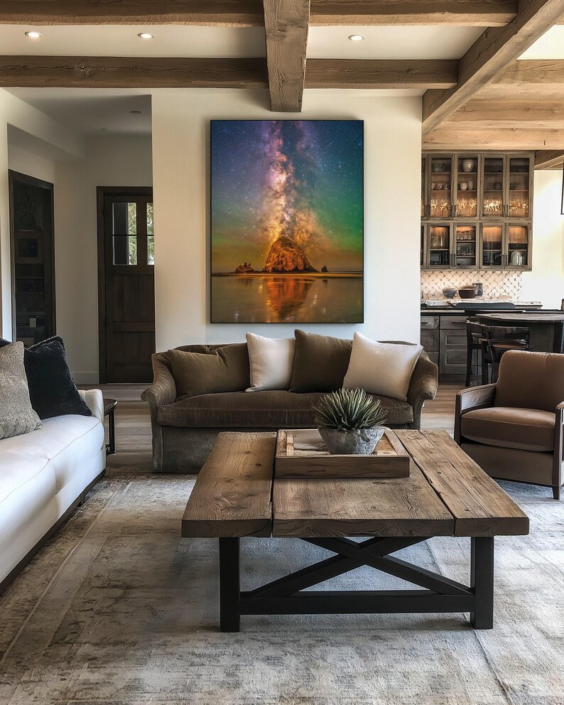May include: A large rectangular wall art print featuring a vibrant night sky with a glowing Milky Way galaxy above a dark coastal rock formation, reflecting onto the wet sand below, displayed in a frame on a white wall above a brown sofa.