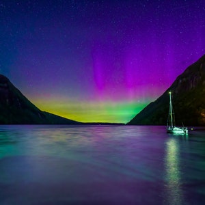 May include: A nighttime landscape featuring a sailboat on a lake under a vibrant display of the aurora borealis. The sky transitions from deep blue to purple and green, with a backdrop of mountains and a small illuminated structure.