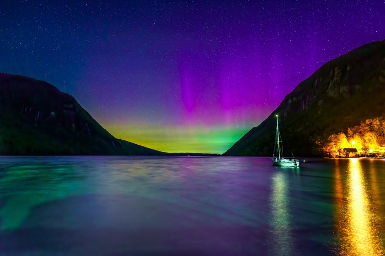 May include: A nighttime landscape featuring a sailboat on a lake under a vibrant display of the aurora borealis. The sky transitions from deep blue to purple and green, with a backdrop of mountains and a small illuminated structure.