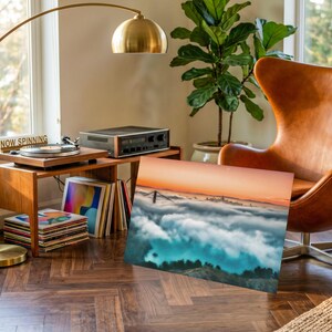 May include: A rectangular wall art print featuring a landscape photograph of a bridge partially obscured by a thick layer of low-hanging clouds at sunrise, with a gradient sky transitioning from orange to blue above the dense white cloud cover.