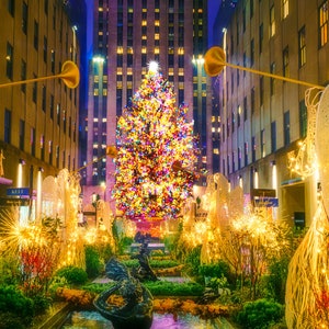 Op de afbeelding: Een grote, fel verlichte kerstboom staat op een stadsplein, omringd door gloeiende witte engelensculpturen en twinkelende lichtjes. De boom is versierd met kleurrijke ornamenten en lichtjes, wat een feestelijke sfeer creëert.