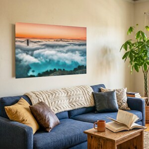 May include: A rectangular wall art print featuring a landscape of the Golden Gate Bridge emerging from a thick layer of fog under a vibrant orange sunset sky. The print is mounted on a light-colored wall above a navy blue sofa with various pillows.