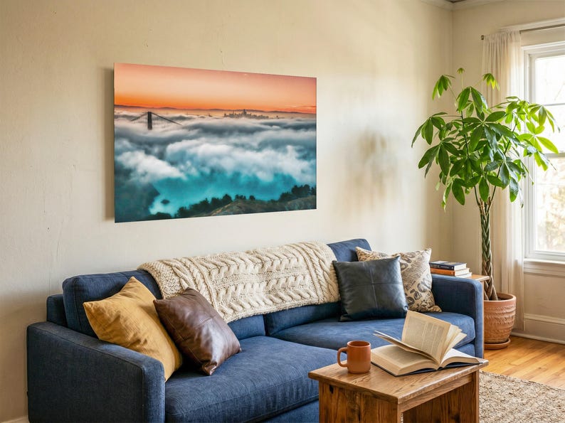 May include: A rectangular wall art print featuring a landscape of the Golden Gate Bridge emerging from a thick layer of fog under a vibrant orange sunset sky. The print is mounted on a light-colored wall above a navy blue sofa with various pillows.
