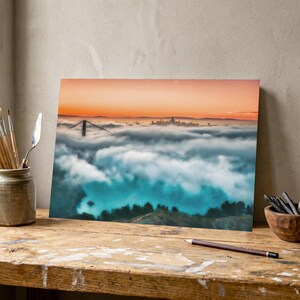 May include: A rectangular print featuring a landscape photograph of the Golden Gate Bridge partially obscured by a thick layer of white fog under a vibrant orange sunset sky, displayed on a wooden desk with art supplies nearby.