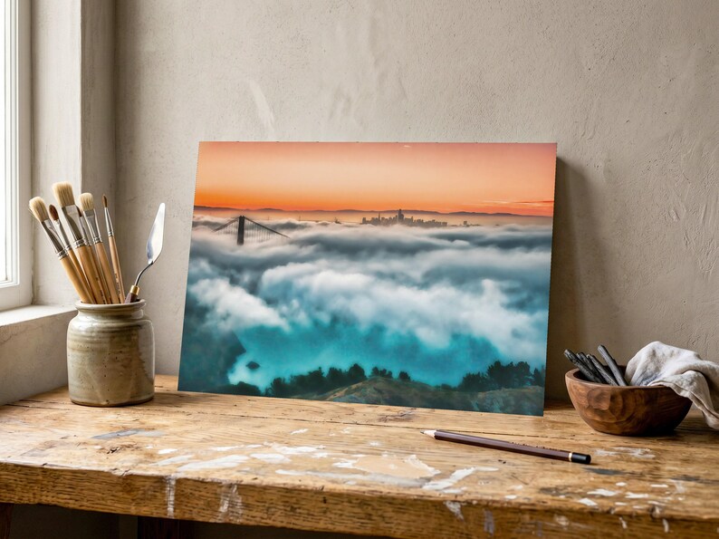 May include: A rectangular print featuring a landscape photograph of the Golden Gate Bridge partially obscured by a thick layer of white fog under a vibrant orange sunset sky, displayed on a wooden desk with art supplies nearby.