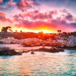 May include: A scenic view of a rocky coastline with a beach and ocean in the foreground. The sky is a vibrant mix of pink, orange, and purple hues, with the sun setting in the distance.