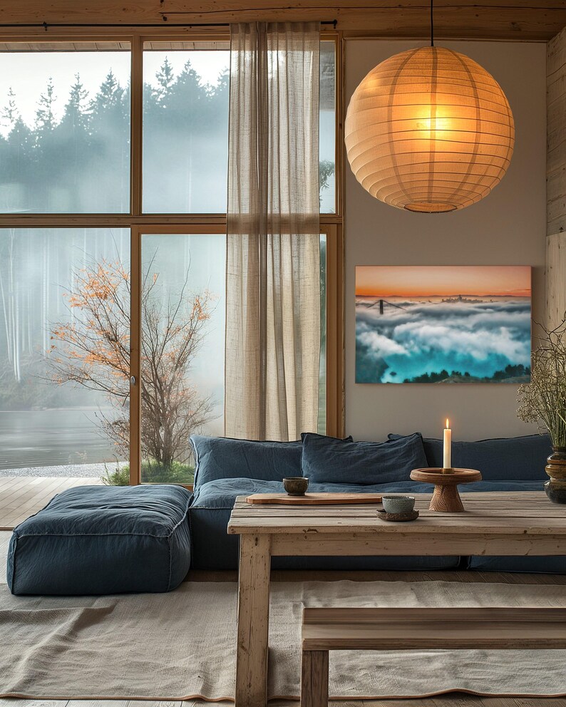 May include: A rectangular wall art print featuring a landscape of a bridge emerging from thick fog under a sunset sky, displayed in a living room with a blue sofa, a wooden table, a lit candle, and a large spherical paper pendant light.