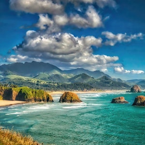 May include: A panoramic view of a rocky coastline with a sandy beach, turquoise water, and a mountain range in the background. The sky is blue with white clouds.