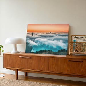 May include: A rectangular wall art print featuring a landscape photograph of the Golden Gate Bridge partially obscured by a thick layer of low-hanging clouds under a vibrant orange sunset sky, displayed as a standalone piece of home decor.