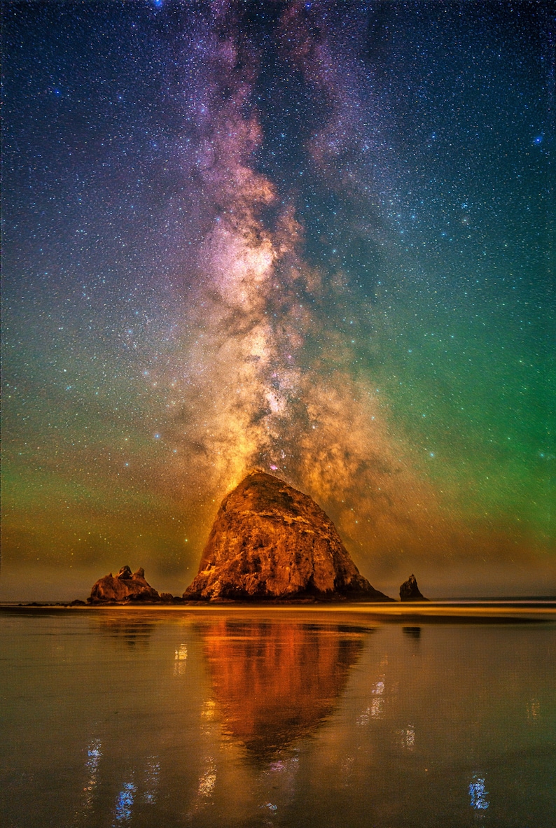 May include: A large, dark, jagged rock formation stands on a wet, reflective beach at night. Above the rock, the vibrant Milky Way galaxy stretches across the sky, displaying hues of deep purple, bright orange, and glowing green starlight.