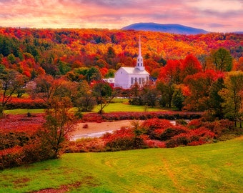 May include: A white church with a steeple stands in a valley surrounded by colorful autumn foliage. The trees are a mix of red, orange, yellow, and green. There is a small stream in the foreground.
